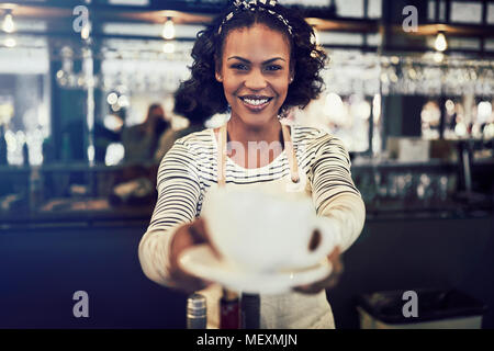 Smiling young African barista seul se tenant derrière le comptoir d'un café branché tenant une tasse de café frais Banque D'Images