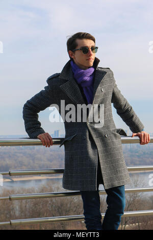 Beau jeune gars dans les lunettes de soleil, foulard et portes manteau près de la main courante par temps ensoleillé. Le coucher du soleil. L'hiver Banque D'Images