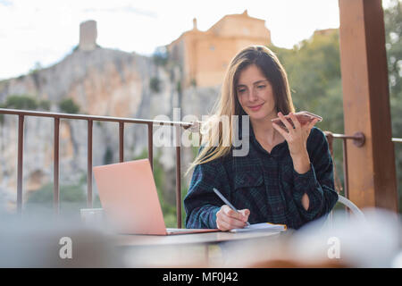 L'Espagne, Alquezar, portrait of smiling young indépendant avec ordinateur portable et téléphone cellulaire travaillant sur terrasse Banque D'Images