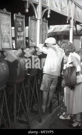 1940, historiques, jeune écolier à l'aide d'un film d'arcade mutoscope viewer lors d'une fête foraine, Londres, Angleterre, Royaume-Uni, avec sa mère à la recherche sur. Ces machines fonctionnant avec des pièces figurant un rabatteur de carte photos encore que fliped donnant ainsi un film d'animation ou film. Banque D'Images