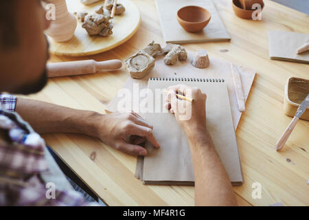 Homme méconnaissable le travail sur le design du céramiste artisanat futur assis à table en bois avec un crayon et un carnet de croquis Banque D'Images