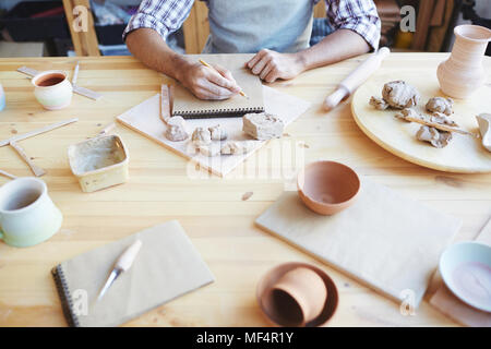 Homme méconnaissable potter dessin dans sketchbook assis à table en désordre avec de l'argile artisanat en atelier Banque D'Images