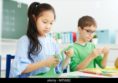 Petite fille asiatique et son camarade de race blanche du papier de couleur avec des ciseaux de coupe tout en faisant la leçon sur l'art de l'origami à l'école Banque D'Images