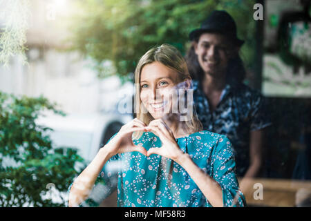 Belle jeune femme faisant un doigt frame heart et souriant à son petit ami au travers d'une fenêtre Banque D'Images