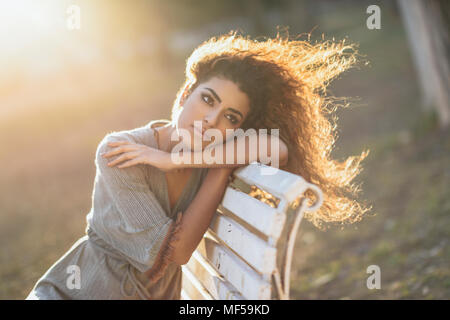 Espagne, Andalousie, Grenade. Belle jeune femme avec curly hairstyle assis sur un banc dans un parc urbain. Concept de vie. Banque D'Images