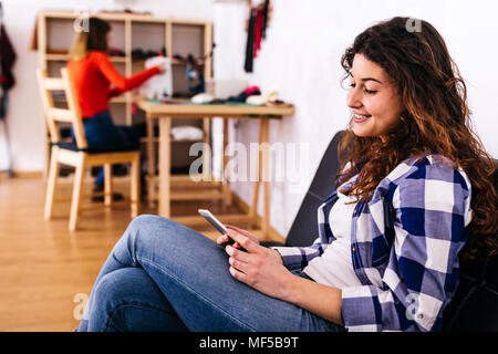 Souriante jeune femme dans mode studio en utilisant un téléphone cellulaire avec femme en arrière-plan sur la machine à coudre Banque D'Images