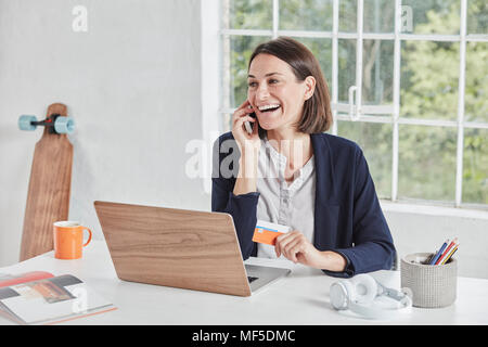 Laughing woman on cell phone at desk holding card Banque D'Images