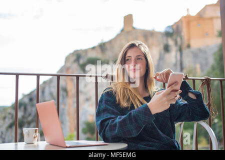 L'Espagne, Alquezar, portrait de jeune femme détendue avec ordinateur portable et téléphone cellulaire assis sur terrasse Banque D'Images