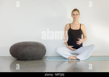 Portrait of smiling pregnant woman sitting sur un tapis de yoga Banque D'Images