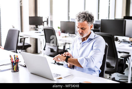 Mature businessman sitting at desk in office looking at smartwatch Banque D'Images