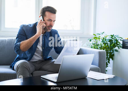 Homme sérieux avec des documents et ordinateur portable sur canapé talking on cell phone Banque D'Images