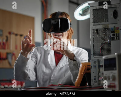 Femme avec des lunettes de VR et manteau de laboratoire assis dans un laboratoire électronique, Autriche Banque D'Images