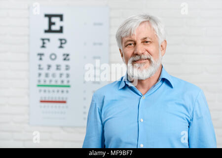 Ancien patient restant en face de l'inspection visuelle tableau accroché sur mur blanc en laboratoire d'ophtalmologie. Homme portant une chemise bleue et une barbe. Essayer d'améliorer la vision de la santé de l'œil. Banque D'Images