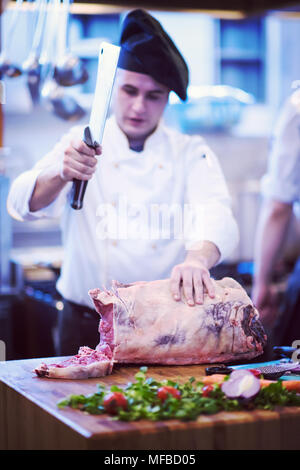 Chef cuisinier avec hache lors de la coupe grand morceau de boeuf sur planche de bois dans une cuisine de restaurant Banque D'Images