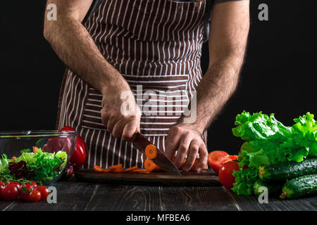 Jeune homme en tablier cook accueil carotte en tranches avec un couteau de cuisine. Banque D'Images