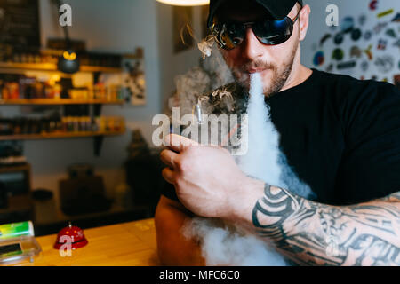 Homme barbu brutale dans une fumée de cigarette électronique lunettes et libère des nuages de vapeur à la vape shop. Banque D'Images