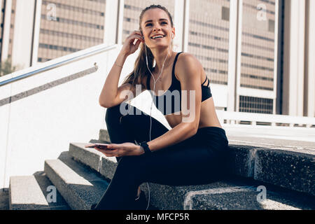 Jeune femme en tenue d'exécution assis sur les marches à l'extérieur et à l'écoute de musique à partir de téléphone mobile. Athlète féminin de prendre de pause après mes rêves Banque D'Images