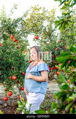 La grossesse et la nutrition - portrait de femme enceinte avec les mains sur son ventre sur le coucher du soleil en arrière-plan le jardin de fruits de grenade. Concept de fertilité. Se Banque D'Images