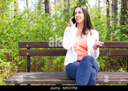 Jolie jeune femme habillée avec son smartphone nonchalamment assis sur un banc en bois dans la forêt. Mode de vie moderne à l'aide de ppc portable Banque D'Images