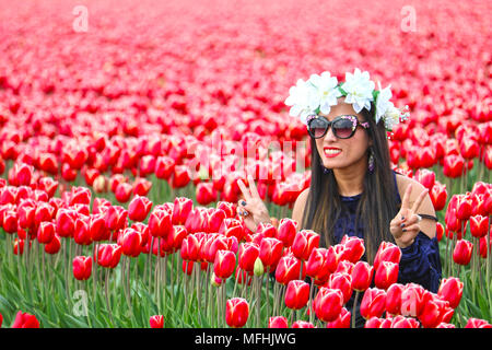 Jeune femme dans un champ de tulipes avec ses doigts vers le haut montrant un signe de paix. Elle porte un bouquet de fleurs blanches sur la tête et lunettes de soleil. Banque D'Images
