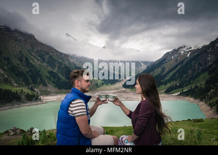 Jeune couple ressemble à l'autre et tient en mains les tasses. Bel homme boit le thé avec petite amie. De majestueuses montagnes aux cimes enneigées, lak Banque D'Images