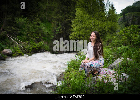 Jeune femme assise sur la pierre dans le parc national et donne sur la rivière de montagne d'ébullition. Girl looking away et souriant sur fond de collines verdoyantes et arbres Banque D'Images