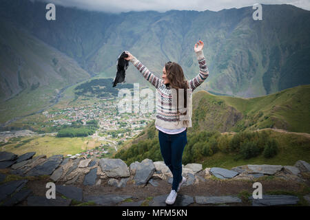 Jeune femme debout sur la colline et d'élever les mains. Jolie fille sur le fond d'une petite ville de la vallée et des montagnes majestueuses. Banque D'Images