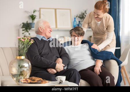 Cheerful senior couple assis sur un canapé avec un fournisseur d'offres debout à côté d'eux et mettre une couverture bleue sur l'épaule de la femme dans un brig Banque D'Images