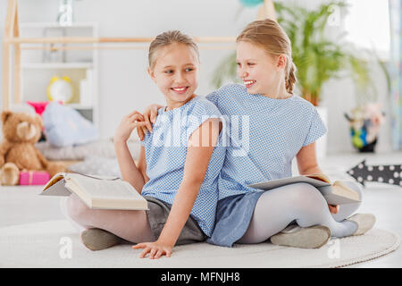 Girl rire avec un bras autour de l'épaule de sa sœur, assise sur un tapis blanc et lire ensemble Banque D'Images