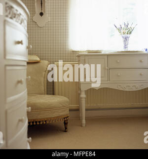 Une chambre à coucher traditionnel blanc détails commode avec un vase de lavande sur le dessus à côté d'un fauteuil rembourré et en dessous d'une fenêtre. Banque D'Images