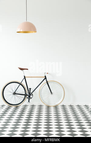 Vélo noir contre mur blanc en intérieur simple avec sol en damier et lampe rose Banque D'Images