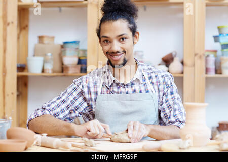 Portrait de jeune beau mâle potter smiling at camera while sitting at table et vase d'argile dans l'atelier de sculpture Banque D'Images