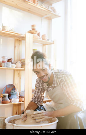 Portrait of young mixed race male céramiste smiling at camera tout en faisant la roue sur les poteries en terre cuite dans l'atelier Banque D'Images