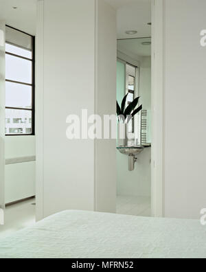 Un détail d'un blanc minimaliste moderne chambre à coucher avec lit double porte ouverte à la salle de bain avec lavabo en acier Banque D'Images