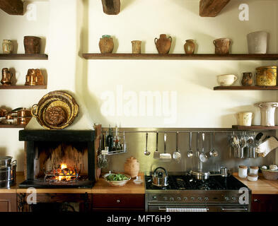 Cuisine de campagne avec un acier inoxydable dosseret, petite cheminée, allumée, et une collection de poterie rustique sur les étagères en bois. Banque D'Images