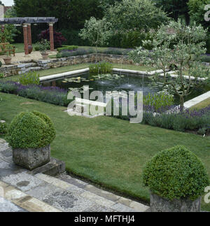 Jardin formel avec une terrasse dallée, étang, murs en pierre, pergola, et plantes en pot. Banque D'Images