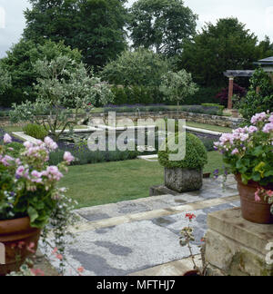 Jardin formel avec une terrasse dallée, étang, murs en pierre, et plantes en pot. Banque D'Images