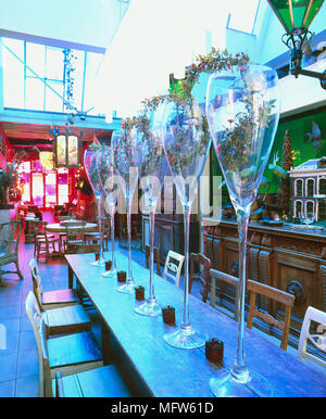 Détails d'une grande salle à manger dans un restaurant au style bohème avec une longue table en bois avec de grands vases en forme de verre à vin de fleurs. Banque D'Images