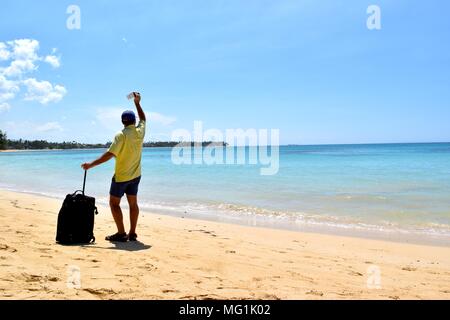 Un homme d'âge moyen sur une plage de sable tenant une valise et un billet dans les mains. Banque D'Images