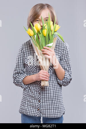 Teen girl hiding face behind bouquet de fleurs sur fond gris. Enfant avec bouquet de tulipes jaunes comme un cadeau. Heureux les mères, anniversaire ou Saint Valentin Banque D'Images