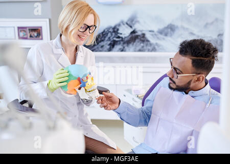 Jeune modèle anatomique stomatologist holding de crâne humain dans les mains tout en discutant avec le traitement de patients de race mixte, soins dentaires prix interio Banque D'Images
