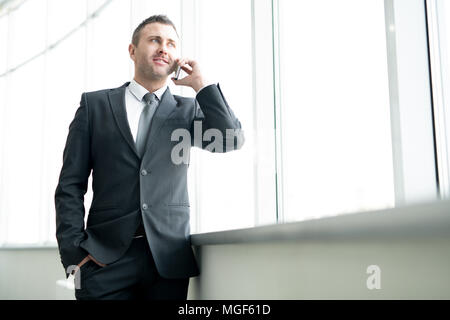 Homme de confiance positif en costume officiel debout à la fenêtre et contempler la ville tout en parlant sur téléphone mobile sur Office Banque D'Images