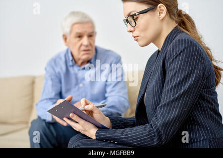 Voir le profil de jeunes psychologue prenant des notes nécessaires tout en ayant rendez-vous au bureau moderne, senior patient Banque D'Images