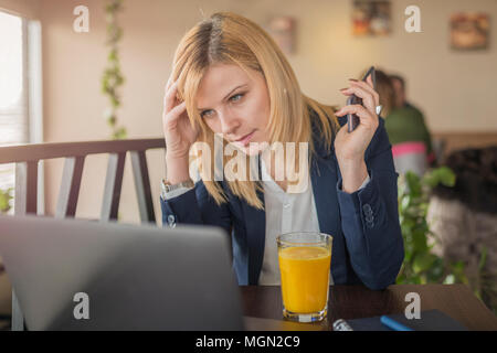 Jeune femme d'affaires nerveux en utilisant son ordinateur portable dans un restaurant. Jolie femme blonde travaille sur son ordinateur portable et l'utilisation de smart phone in Banque D'Images