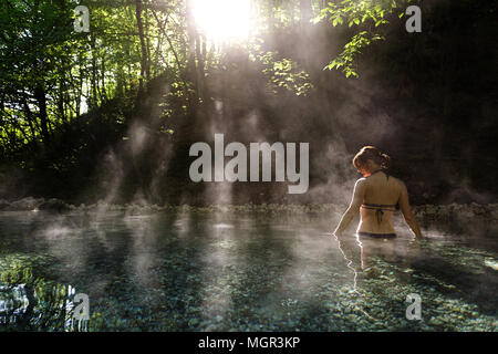 Young woman in bikini debout dans une piscine naturelle d'eau chaude dans la forêt avec rétro-éclairage et la vapeur autour d'elle, Maibachl, Autriche. Banque D'Images