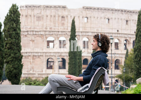 Beau jeune homme en chiffons sport écouter de la musique assis sur un banc avec le Colisée en arrière-plan. Les jeunes sportifs en survêtement 23-08-2003 détente Banque D'Images