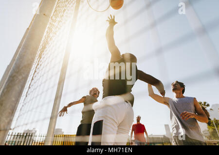 Quatre hommes jouant au basket-ball dans une cour de basket-ball lors d'une journée ensoleillée. Man jumping high de lancer la balle dans le panier. Banque D'Images