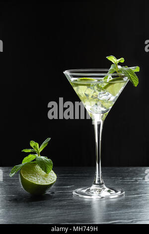 Fraîcheur d'un cocktail avec de la glace concassée, tranches de lime et de feuilles de menthe dans un verre à martini sur fond noir rétroéclairé Banque D'Images