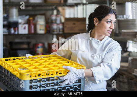 Une femme travaillant dans une cuisine commerciale. Elle porte un uniforme blanc et des gants en latex, transportant une caisse en plastique. Banque D'Images