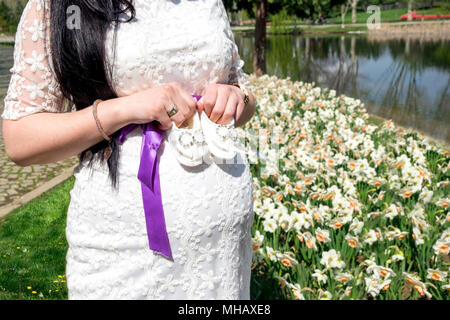 La femme enceinte attend son bébé. Cette photographie prend alors qu'elle tenait des chaussures dans sa main devant des fleurs et un lac. Banque D'Images